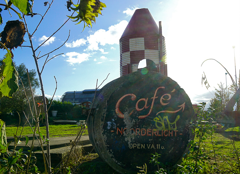 Auf einer Wiese in Amsterdam steht ein schwarzes, rundes Schild des Cafés Noordlicht, dahinter ein kleiner Leuchtturm in rot-weiß aus Blech.