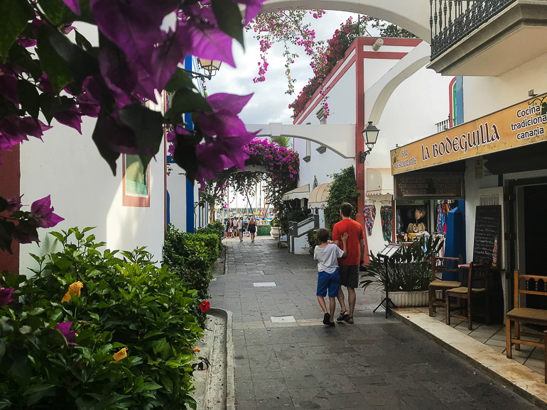 Unter lila Blütenbögen und an kleinen Tavernen vorbei schlendern Touristen in Las Palmas Fußgängerzone Puerto de Mogan entlang.