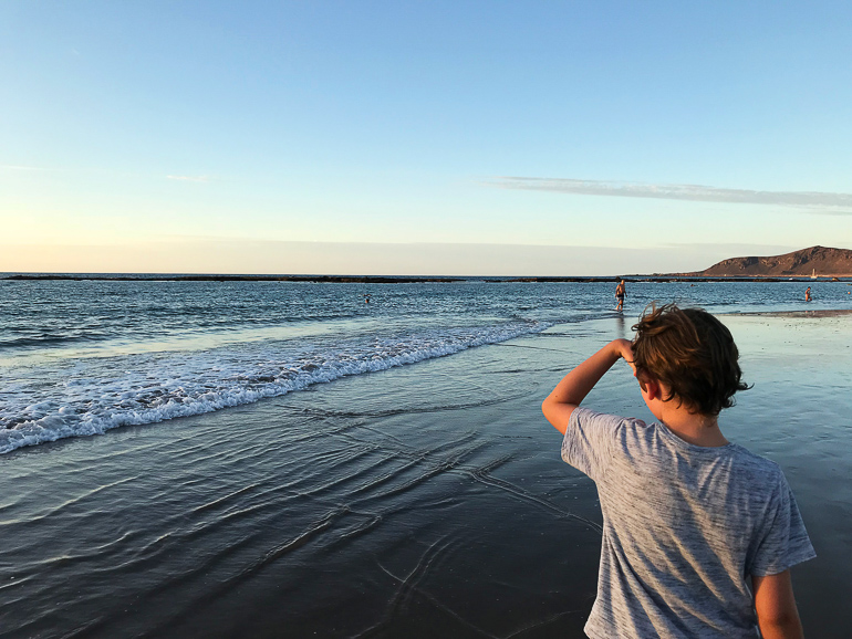 Ein kleiner Junge mit grauem Shirt blickt am Strand von Playa de Las Canteras auf das Meer.
