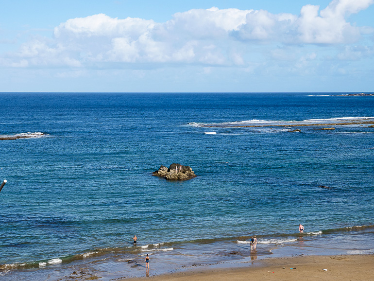 Am Sandstrand bei Las Palmas stehen vereinzelt Badegäste am Meer und beobachten die brechenden Wellen an kleinen Felsvorsprüngen.
