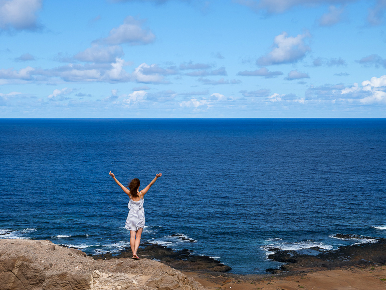 Auf Las Palmas Halbinsel La Isleta steht eine Frau vor dem Meer auf einem Felsvorsprung, sie trägt ein gestreiftes Kleid und streckt beide Arme in den Himmel.