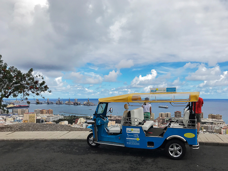 Vor der Kulisse der Uferpromenade hat auf einer Straße in Las Palmas ein blau-gelbes, elektrisches Tuk-Tuk geparkt.