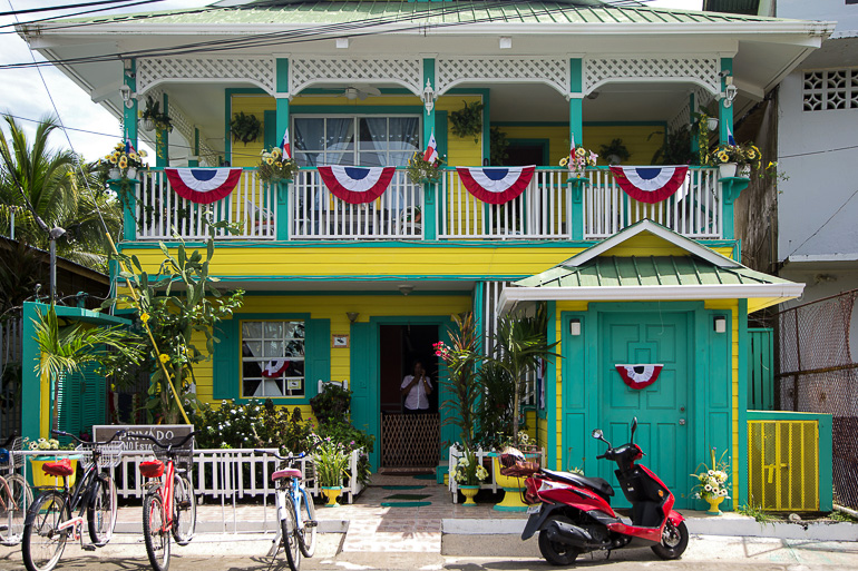 A red scooter is parked in front of a yellow and green colonial-style house richly decorated with plants and flags in Bocas del Toro, Panama.