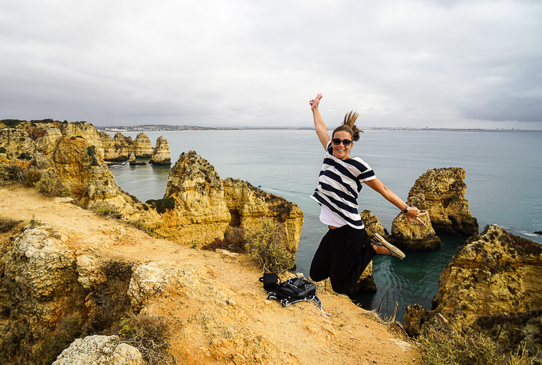 Eine junge Frau mit braunen Haaren, Sonnenbrille und in schwarz-weiß gekleidet hüpft an einer Erhöhung vor dem Meer der Algarve, dem Lagos Ponta Piedade, in die Luft.