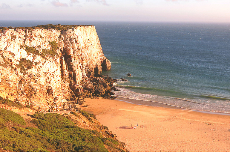 An einem Strand an Portugals Algarve ragt ein riesiger Felsen aus dem Meer, während vereinzelt Menschen am Strand entlang spazieren, die wie Ameisen von einer erhöhten Aussicht wirken.