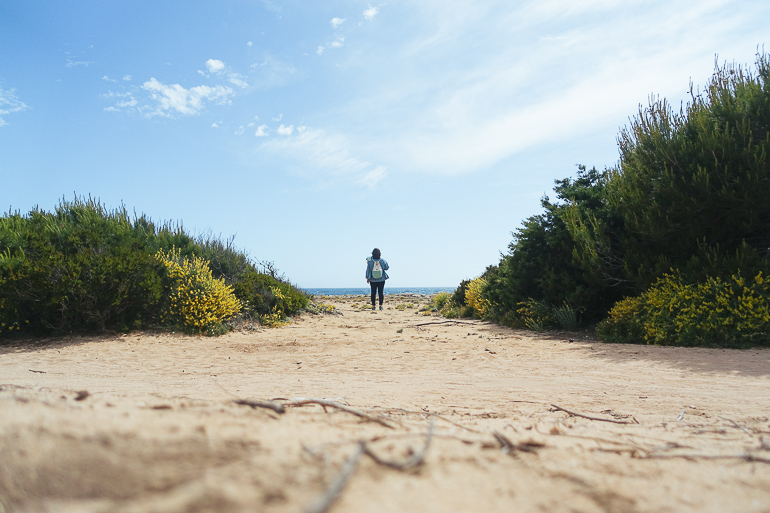 Auf einem sandigen Weg in Mallorca, umgeben von Gebüsch, steht eine Frau mit Rucksack mit Blick aufs Meer.