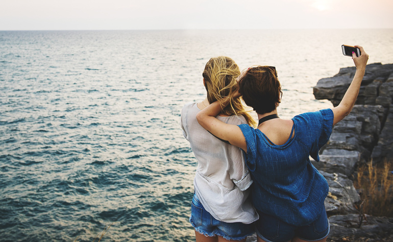 Zwei junge Frauen stehen zueinander geneigt an einer Küste am Meer und machen ein Selfie von sich.