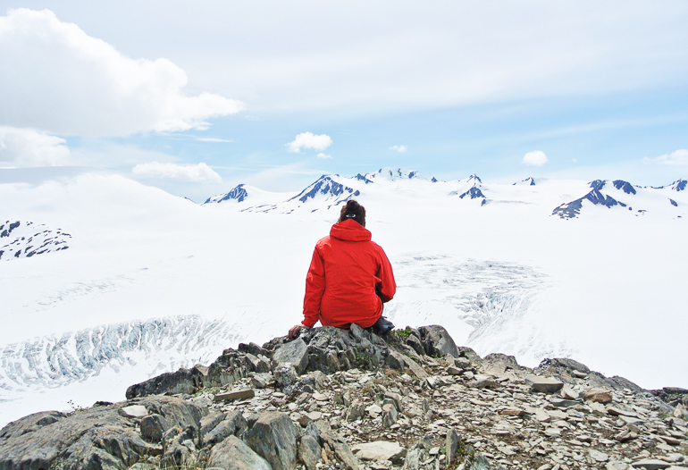 A hiker in a red jacket and black beanie sits on a rocky outcrop looking at the snow-covered glacial landscape of Harding Icefield, Alaska.