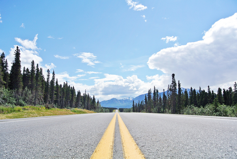 A deserted highway with a backdrop of mountains in Alaska is surrounded by forests.