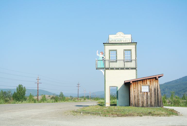 A tall wooden sign in the shape of a western parlor stands on a deserted Alaskan road pointing the way to Dawson City.