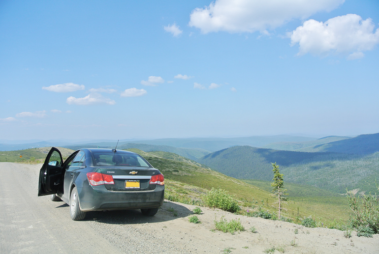 A black Chevrolet with the driver's door open is parked in front of a mountain landscape on Alaska's Top of the World Highway.