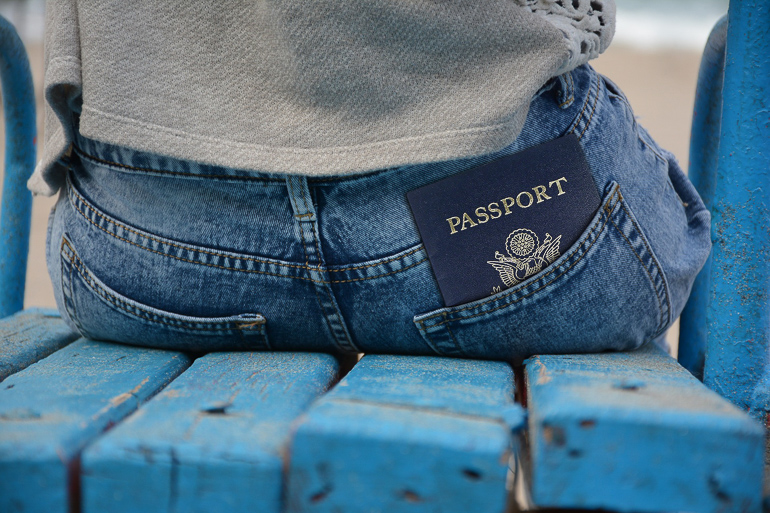 In der Jeans-Gesäßtasche einer Frau, die auf einer blauen Holzbank sitzt, blickt ihr blauer Pass hervor.