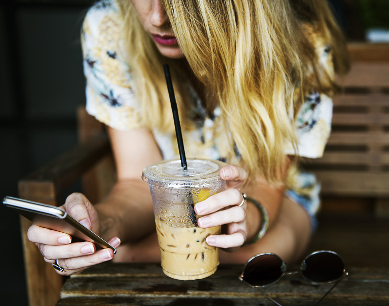 Eine blonde Frau sitzt auf einer Holzbank, und blickt auf ihre Handy in der linken Hand um eine PayPal-Zahlung zu veranlassen, in der rechten Hand hält sie einen Coffee-to-go-Becher.