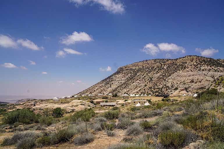 Blick auf das begrünte und kleinen Zelten versehenen Dana Nature Reserve in Jordanien, im Hintergrund ragt ein kleiner Berg in den fast wolkenfreien Himmel.