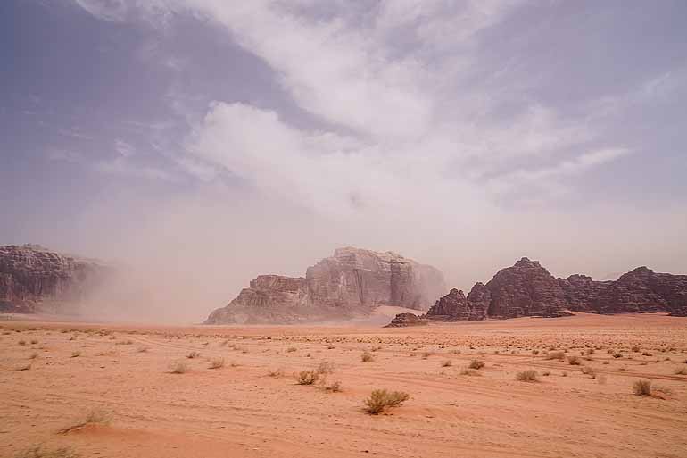 Der Wind wirbelt Sand durch die Wüste Wadi Rum, im Hintergrund ragen Felswände hervor.