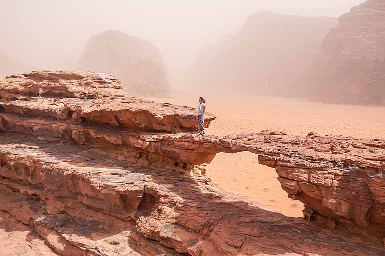 Eine Frau steht in der Wüste Wadi Rum, auf einer Felsformation und blickt auf die unendlichen Weiten.