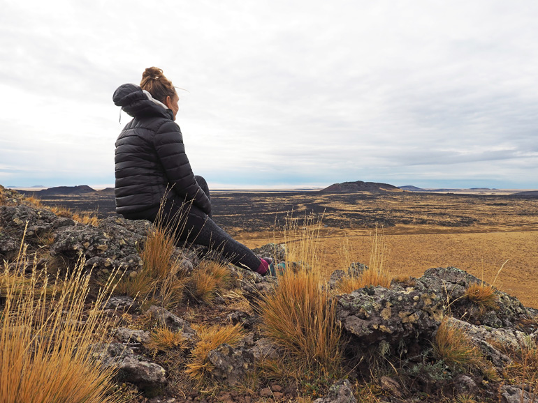 Eine Frau in schwarzer Jacke sitzt auf einem Stein im Pali Aike Nationalpark in Patagonien und blickt in die Ferne.
