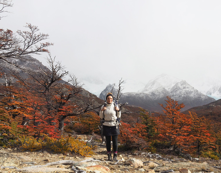 A hiker stands in Los Glaciares National Park between reddish bushes with the mountains of Patagonia in the background, the peaks of which are covered with snow.