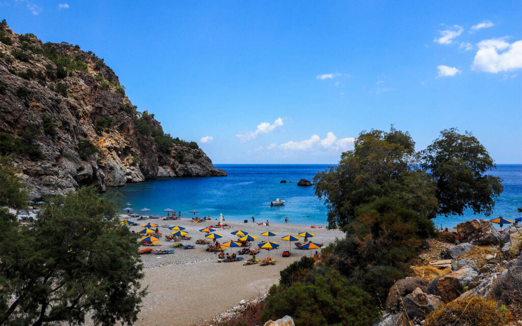 In Karpathos befindet sich zwischen Büschen und Kiefernwäldern am Wegesrand der Acháta-Strand, dessen gelb-blaue Sonnenschirme seine Gäste vor der Sonne schützt.