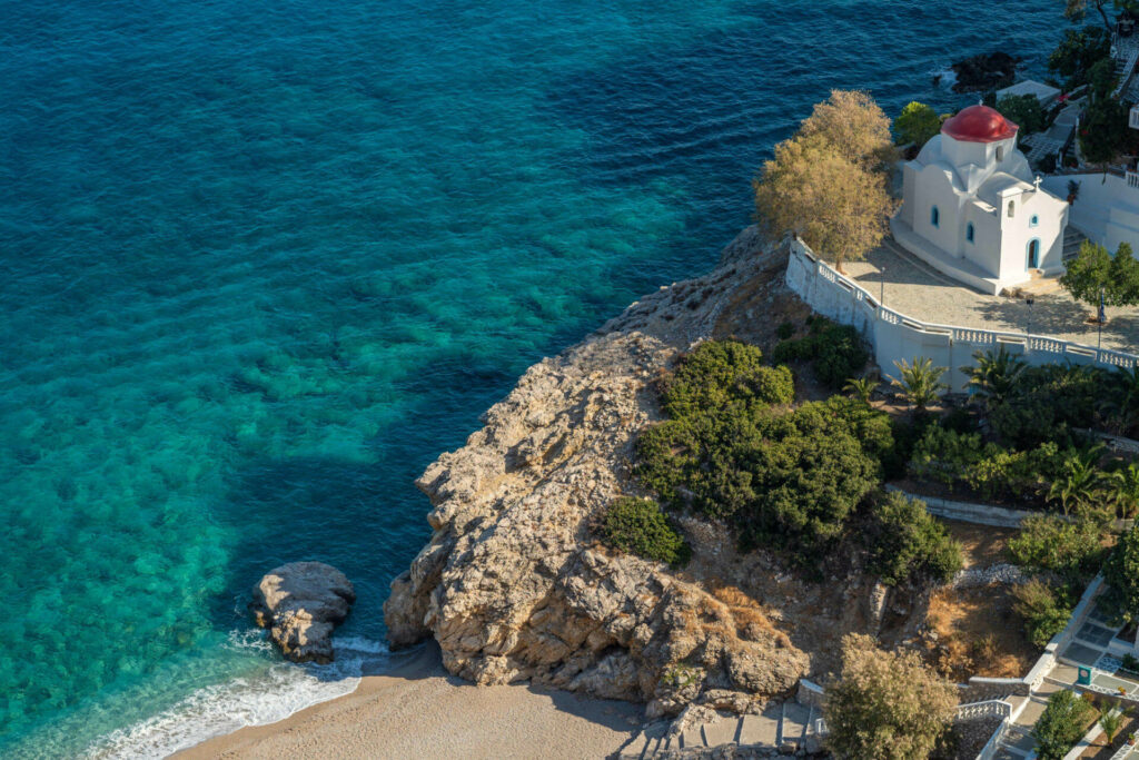 Der Kyra Panagia Strand auf Karpathos liegt idyllisch unter einem Felsen, auf dem ein weiß-rotes kubisches Haus steht.