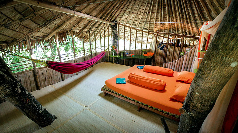 In dem Inneren des Baumhauses von Bukat Raya auf Borneo steht ein Bett aus Holz und orangen Bezügen sowie eine lila Hängematte.