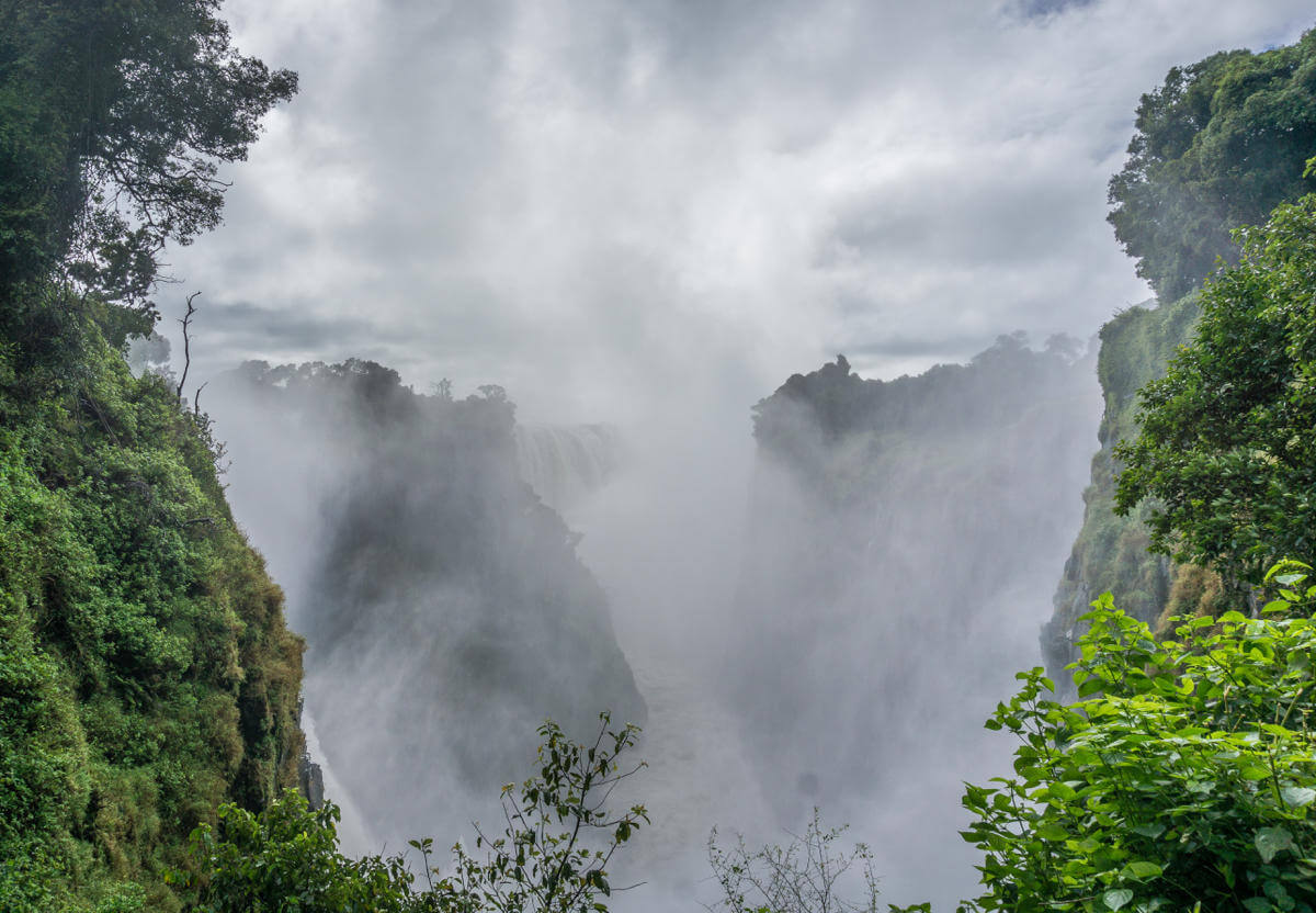 An den Victoriafalls steigt zwischen der üppigen Vegetation Sprühnebel des Wasserfalls in die Luft.