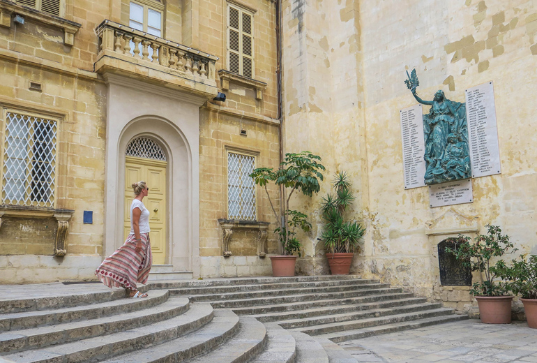 Frau blickt auf Denkmal an Hauswand in Vittorosia, eine der „Drei Städte“