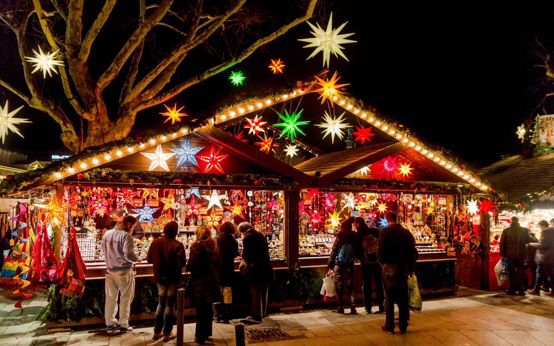 Bunt beleuchteter Stand auf einem Weihnachtsmarkt mit Sternen im Baum und Menschen davor.