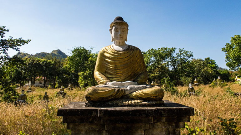In Myanmars Hpa An steht eine sitzenden Buddha-Statue inmitten von hohen Gräsern und Bäumen im Lumbini Buddha Garden.