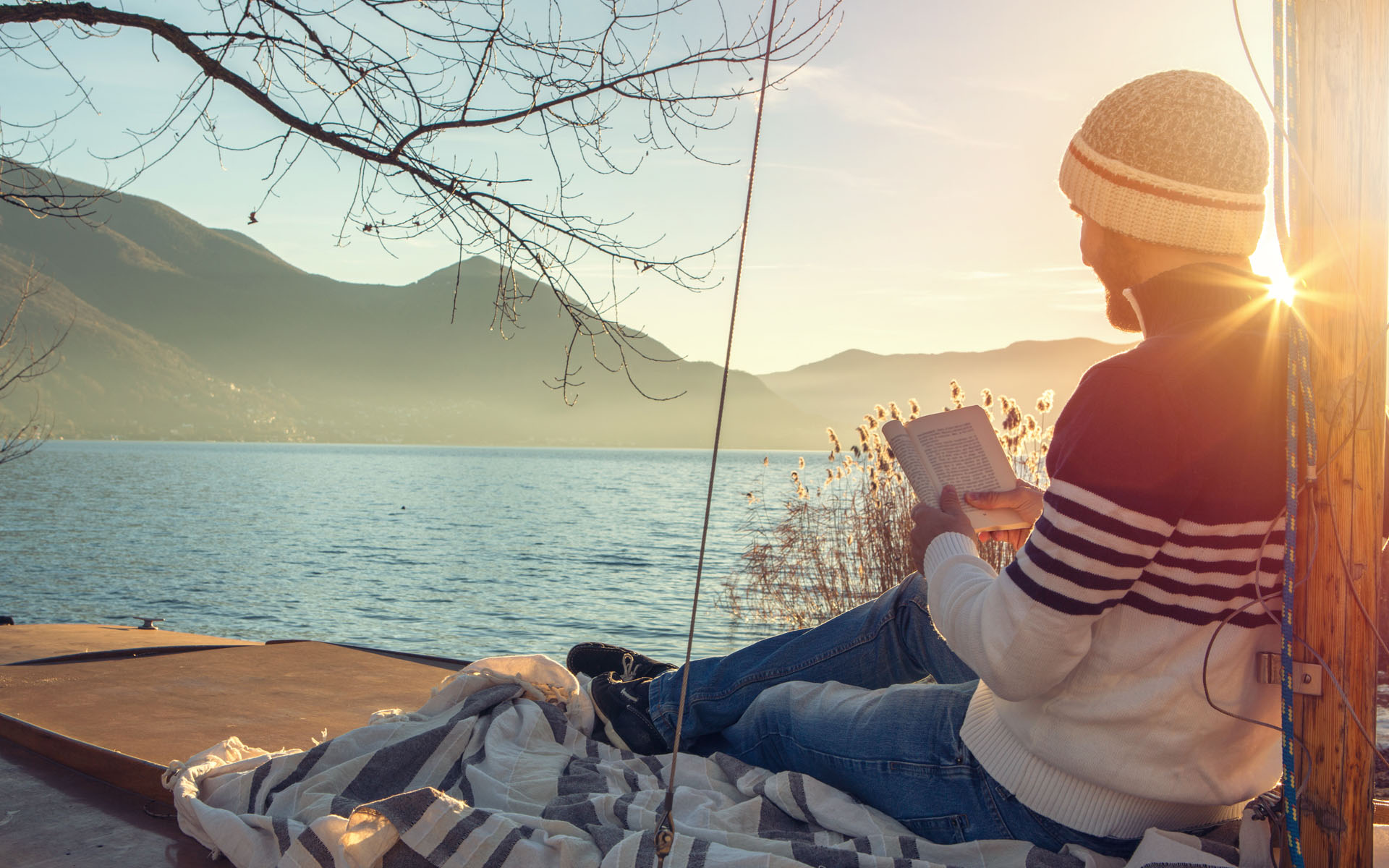 Ein junger Mann sitzt an einem See auf einem hölzernen Segelboot und liest ein Buch.