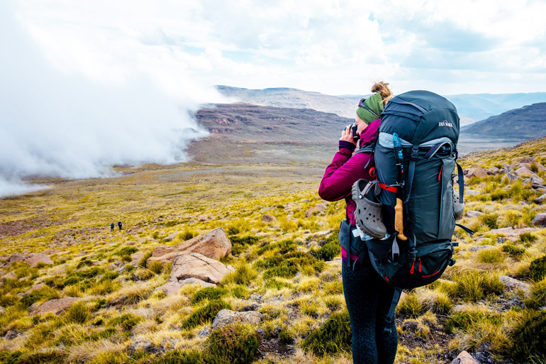 Südafrika Drakensberge: Eine Frau mit einem Rucksack in einem Tal fotografiert die Landschaft.