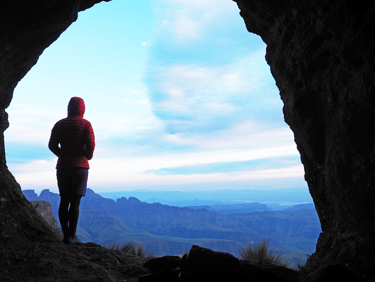 Südafrika Drakensberge: Eine Frau steht in einer Höhle und blickt auf die Landschaft.