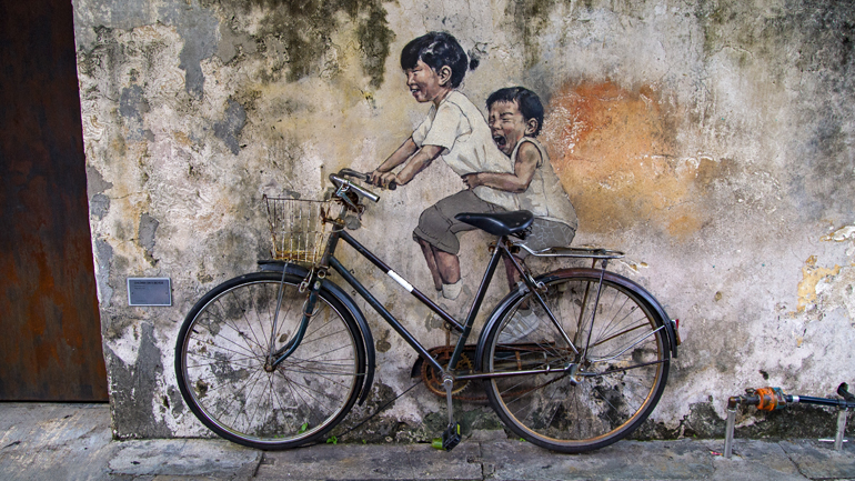 Malaysia: Zwei gezeichnete Kinder sitzen auf einem echten Fahrrad.