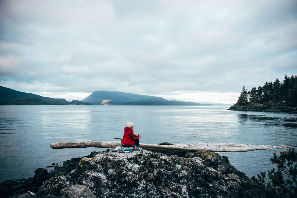 Vancouver Island: Eine Frau mit einer roten Jacke, sitzt auf einem Felsen mit Blick auf das Meer.