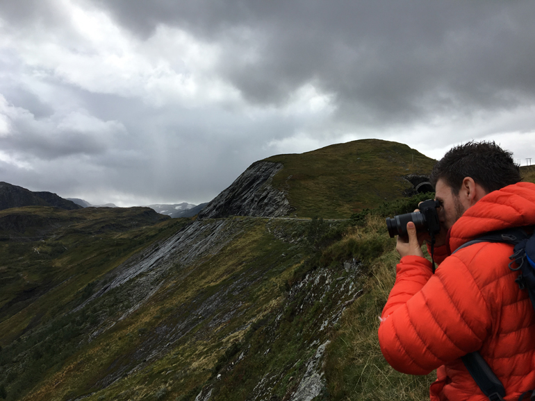 Beste Reisekamera: Ein Mann mit einer Roten Jacke hält eine Kamera in der Hand im Hintergrund sind Berge zu sehen.