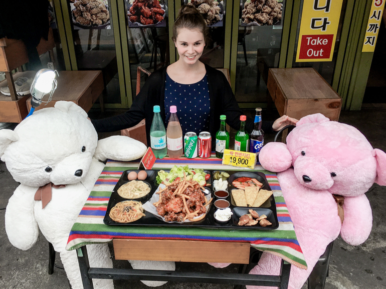 Seoul Sehenswürdigkeiten: Eine Frau sitzt an einem Tisch, vor hier befindet sich allerlei Essen, neben ihr sitzen ein weißer und ein rosafarbener Teddybär.