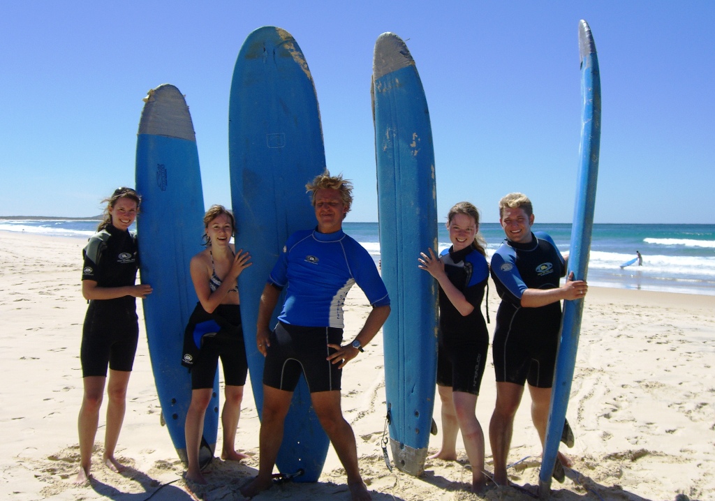 In einem Surfcamp stehen fünf jungen Menschen mit Surfbrettern beieinander am Strand.