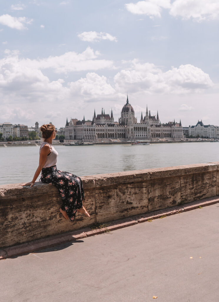 Budapest Städtetrip: Eine Frau sitz auf einer Mauer im Hintergrund ist Wasser zu sehen und ein Gebäude.