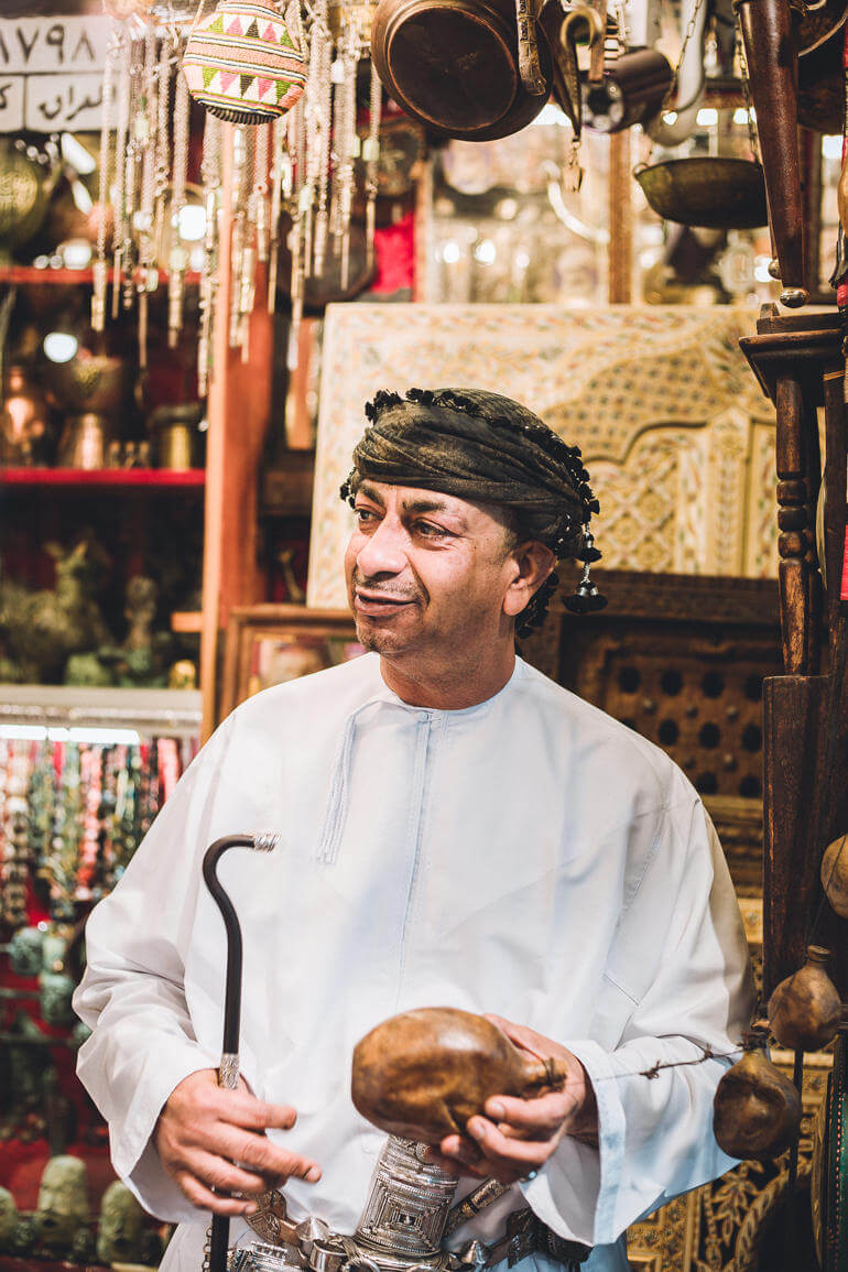 Roadtrip durch den Oman: Ein Mann mit Kopfbedeckung in einem Basar Geschäft hält einen Stock in der Hand.