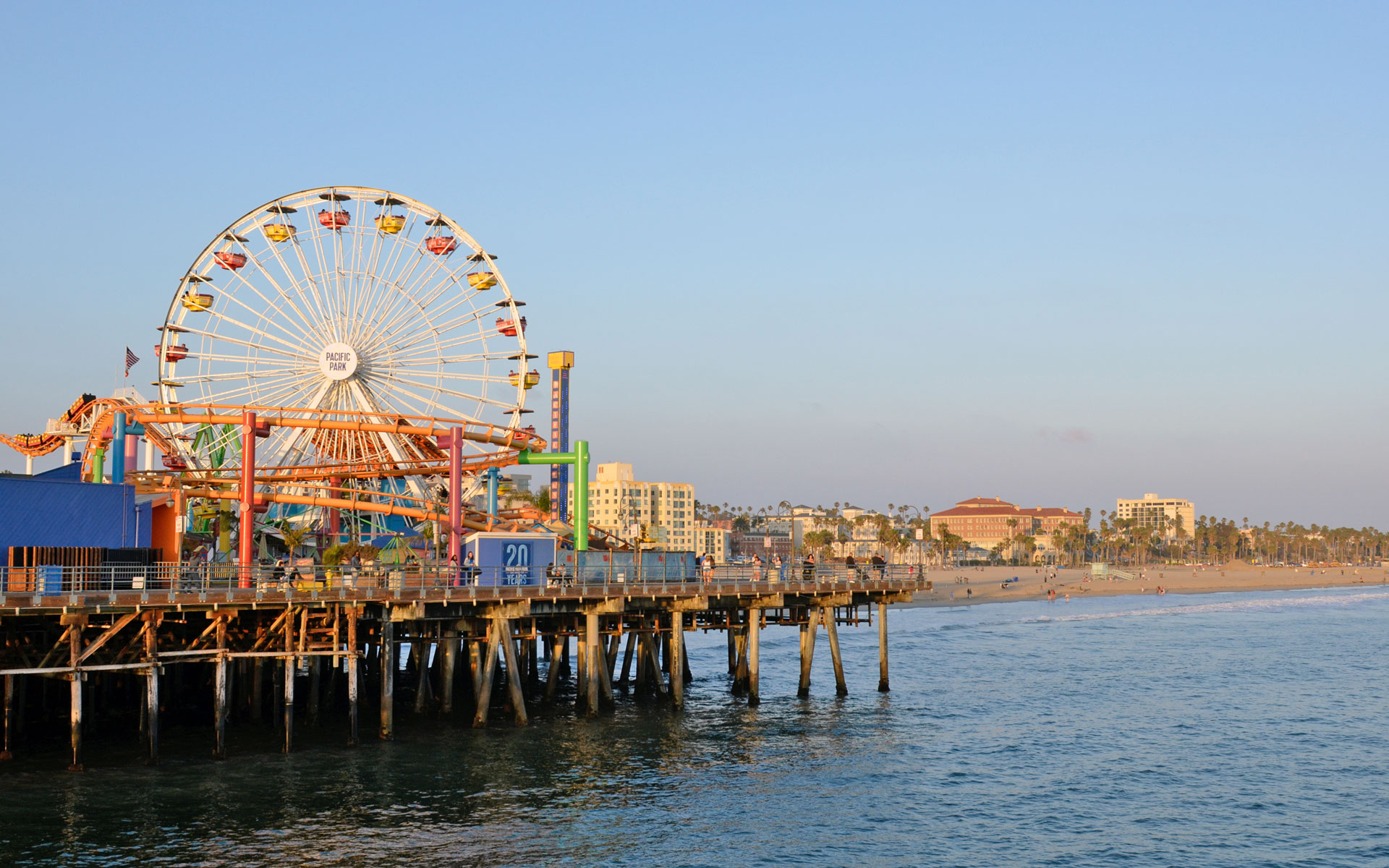Fahrgeschäfte, darunter ein Riesenrad, auf dem berühmten Santa Monica Pier in der Nähe von Los Angeles, Kalifornien. Im Hintergrund leuchtet der Strand im Sonnenuntergang.