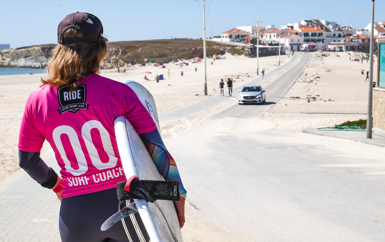Eine Frau mit einem Surfbrett von hinten geht einen Weg entlang in Peniche, Portugal.