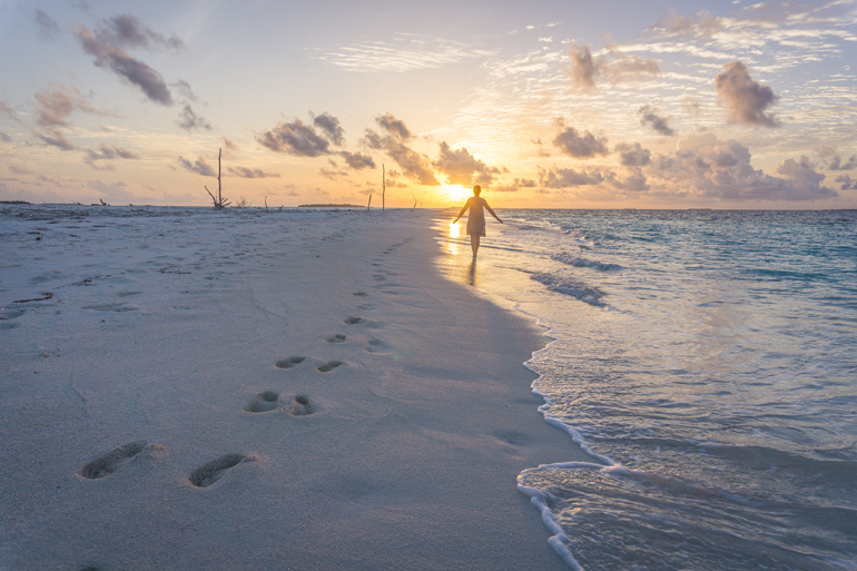 Eine Frau läuft am Strand entlang dem Sonnuntergang und hinterlässt Fußspuren im Sand.
