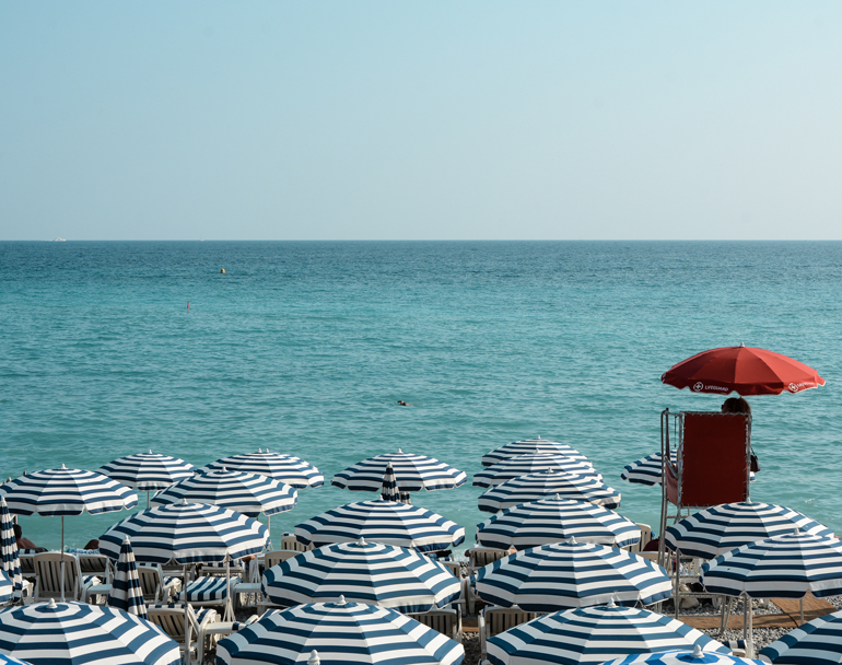 Das Blau der gestreiften Sonnenschirme wetteifert mit dem von Himmel und Meer. Blau - weiße Sonnenschirme stehen am Strand.