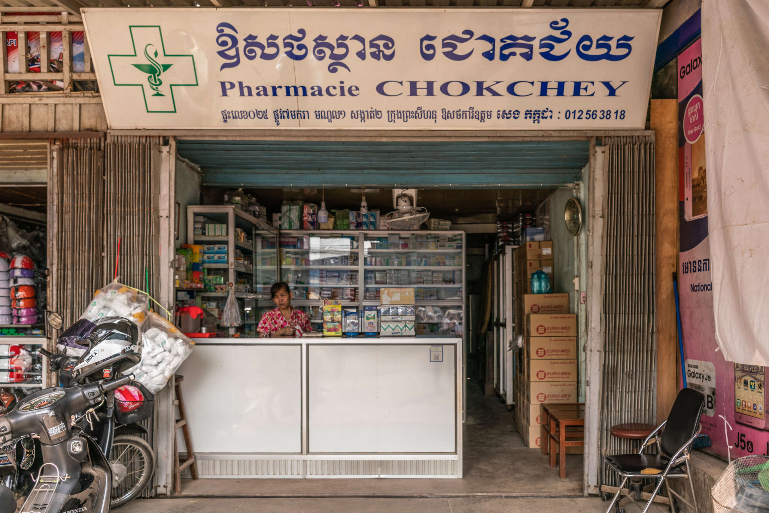 Eine Frau steht hinter einem Apothekentresen eines offenen Geschäfts mit Apothekenzeichen und der Aufschrift "Pharmacie Chokchey"