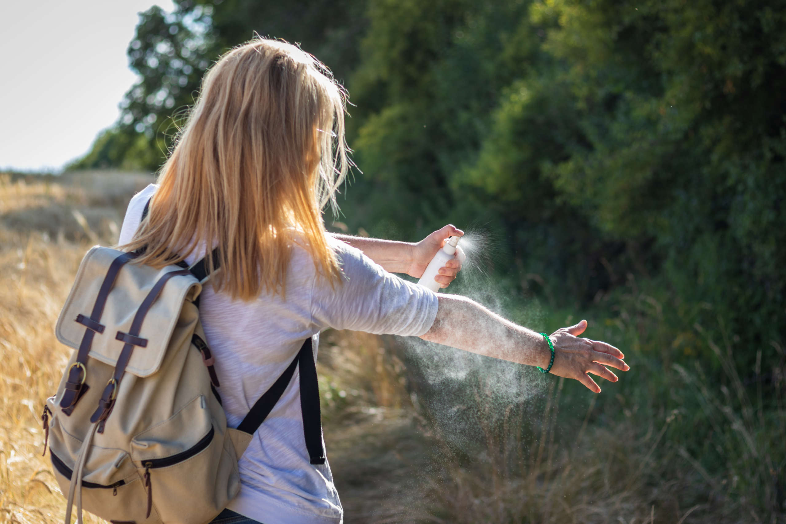 Eine Frau besprüht ihren Arm mit Insektenspray, ihre blonden Haare und beigen Rucksack sieht man von hinten, vor ihr liegen Wald und Feld.