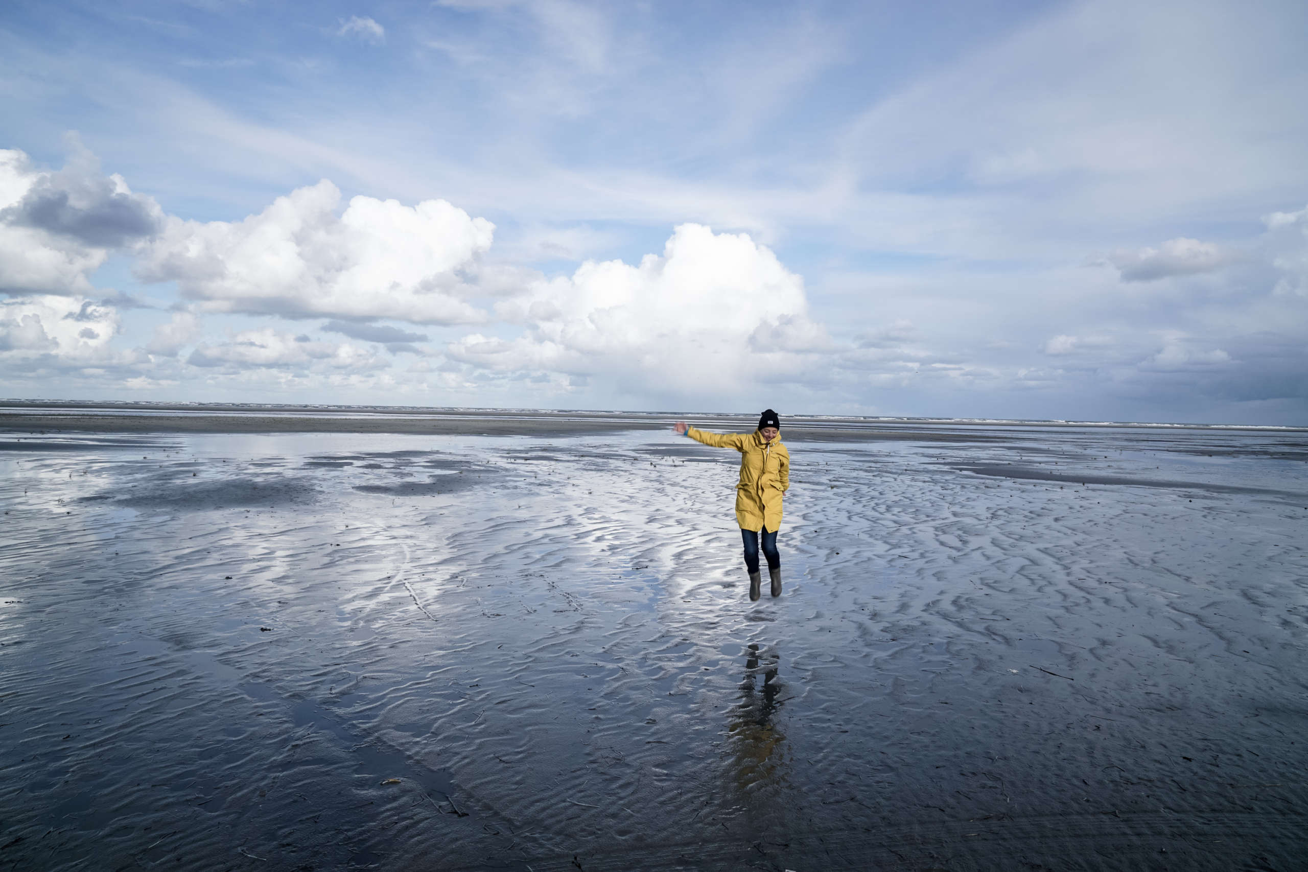Eine Frau im gelben Regenmantel steht bei Ebbe auf dem Wasser auf der Insel Ammeland in den Niederlanden und streckt den Arm heraus