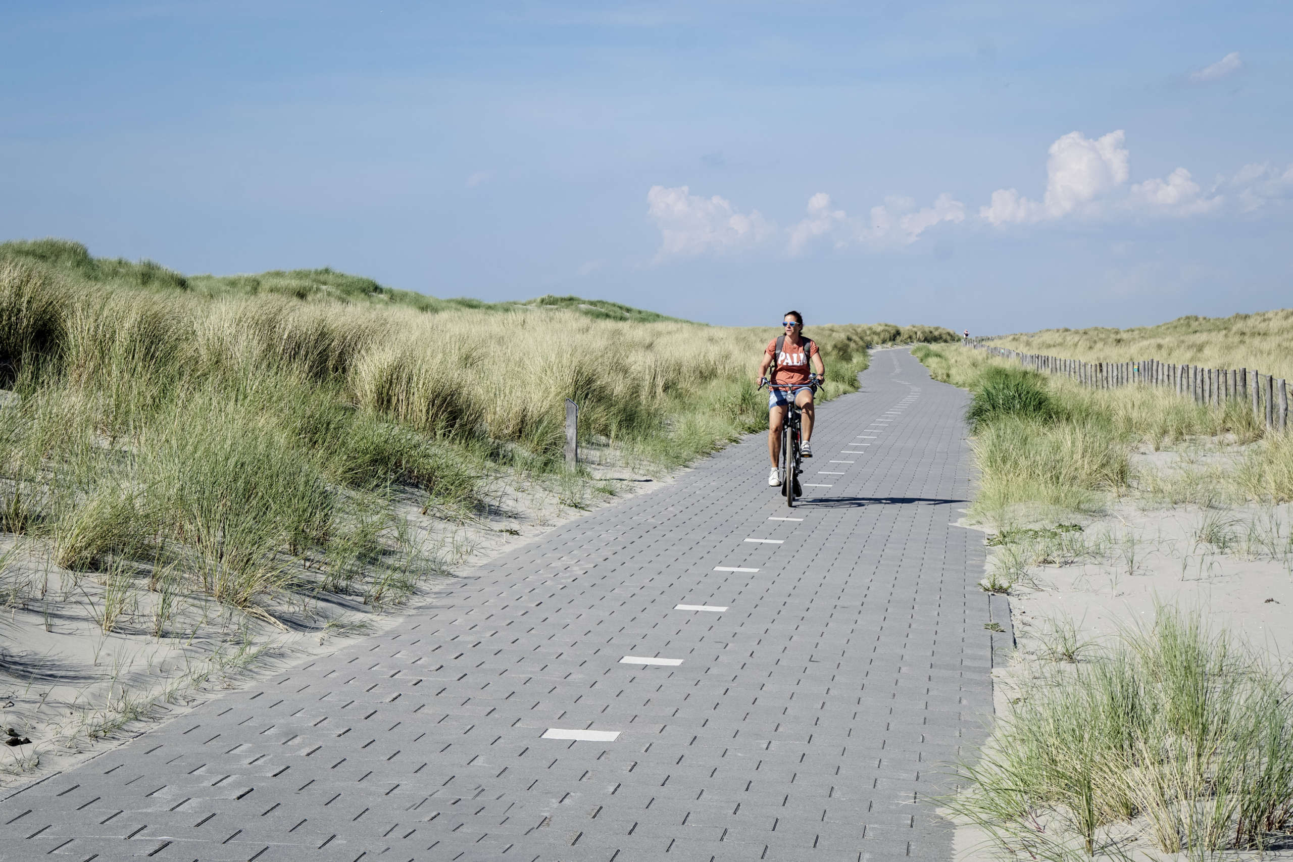 Eine Frau fährt auf einem Fahrradweg Rad, rechts und links von ihr befinden sich Sandanhäufungen auf denen Gestrüpp wächst