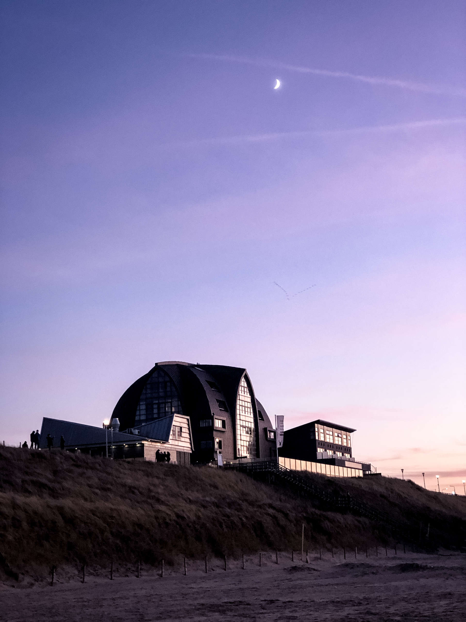 Blick vom Strand aus auf ein architektonisch außergewöhnliches Hotel in den Niederlanden, dahinter der lila gefärbte Himmel, der Halbmond ist zu sehen.