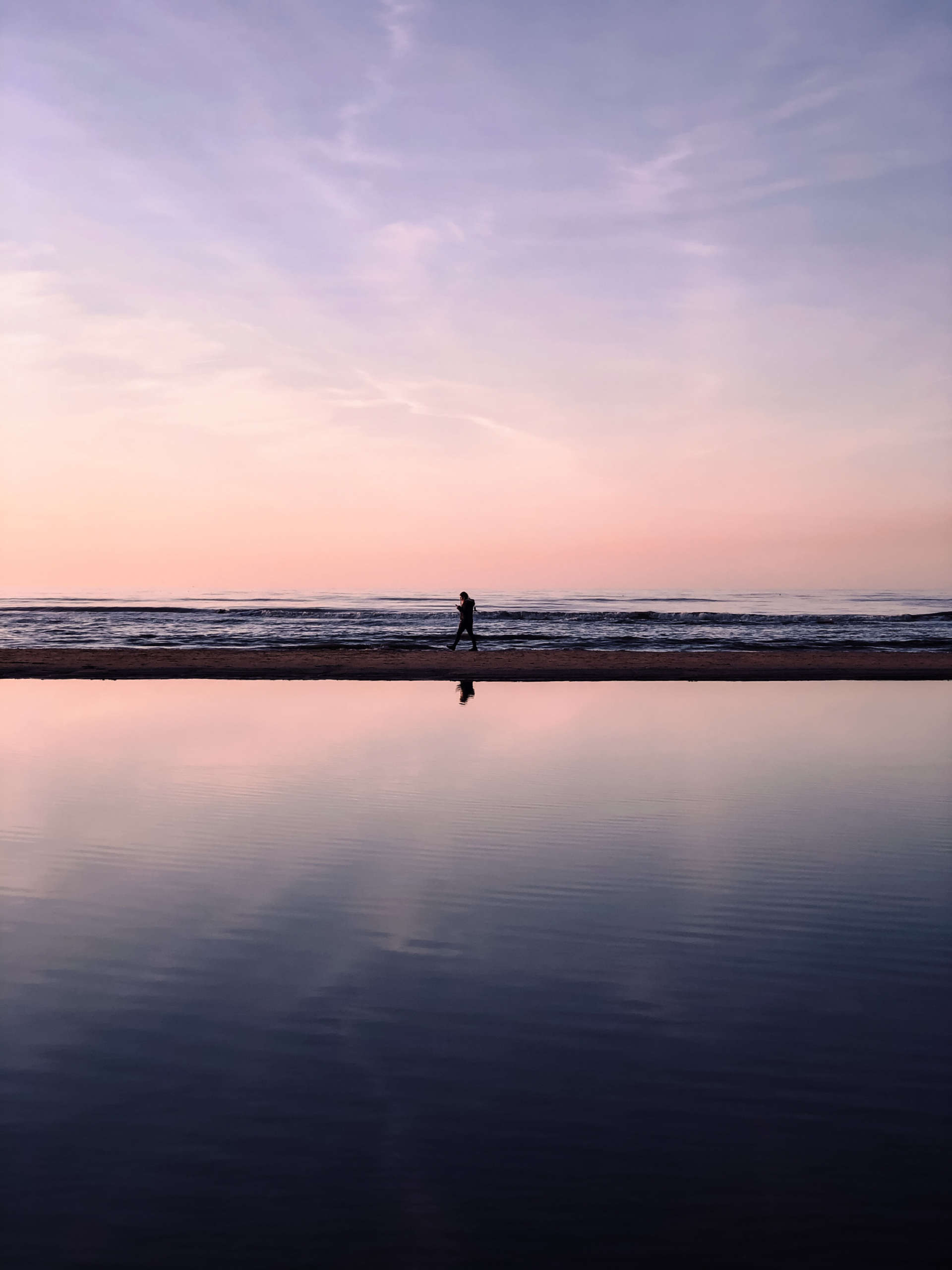 Lila und pink gefärbter Himmel, darunter das Meer und eine kaum erkennbare Person, die den Strand entlang spaziert