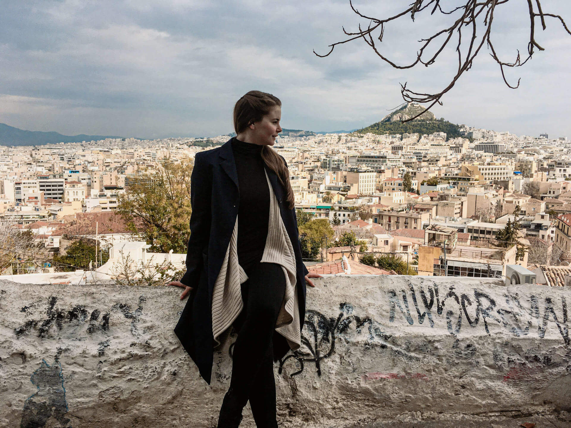 Blick auf die sich weit erstreckende Hauptstadt Athen und die Akropolis von Anafiotika aus, Reisebloggerin Franziska posiert vor der Dächerflut auf einer Mauer sitzend.